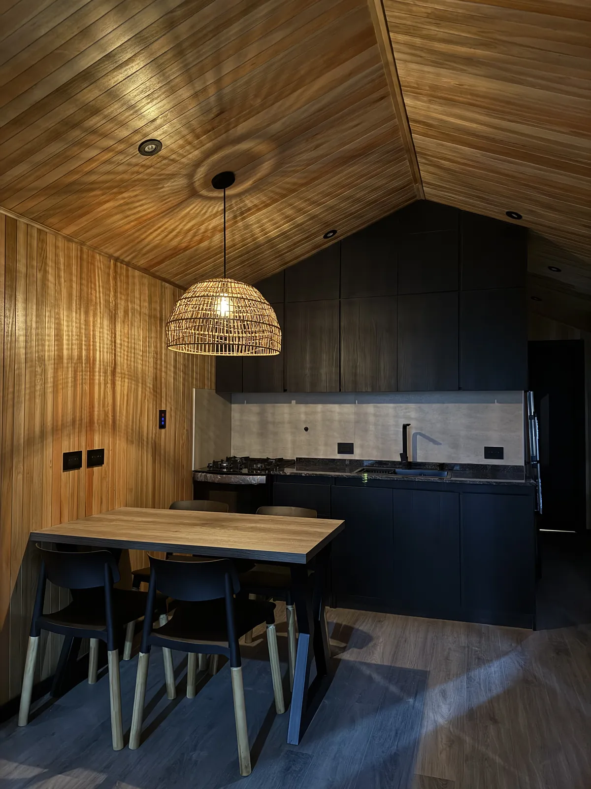 Comedor con lámpara de ratán y cocina negra bajo techo de madera casa modular Faci House