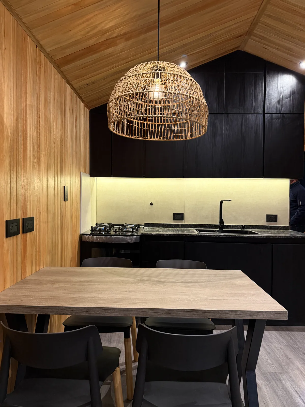 Mesa de comedor frente a la cocina con alacenas negras y luz LED casa modular Faci House