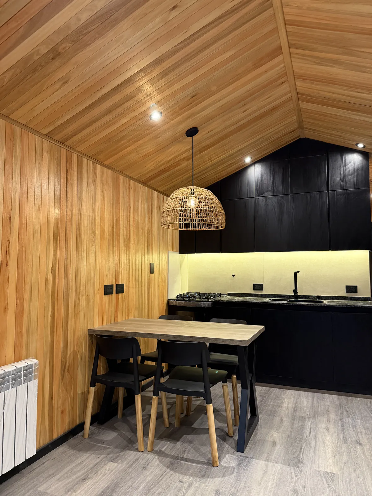 Cocina negra integrada al comedor con lámpara de ratán y techo de madera a dos aguas casa modular Faci House